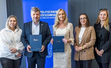 Od lewej prof. A. Sikora, rektor, prof., P. Koszelnik, dr. B. Szafrańska, p. P. Multan, arch. D. Bober. Na zdjęciu na tle ciemnej ściany z wyróżniającym się niebieskim banerem z białym napisem Politechnika Rzeszowska stoi pięć elegancko ubranych osób. Dwie pośrodku trzymają okładki z umowami.