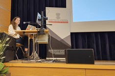 Na zdjęciu widzimy kobietę z długimi, falowanymi włosami, ubraną w jasną koszulę i jeansy, która siedzi przy biurku na scenie i prowadzi prezentację. Przed nią znajduje się laptop oraz sprzęt multimedialny, w tym mikrofon i monitor. Obok niej stoi baner. Na pierwszym planie widoczne są również ozdobne rośliny doniczkowe. Tło stanowi ciemna zasłona sceniczna i fragment dużego ekranu projekcyjnego.