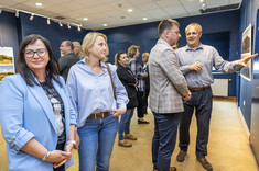 W pomieszczeniu na pierwszym planie stoją dwie kobiety. Obie ubrane na niebiesko. Jedna uśmiecha się do zdjęcia. W tle dwóch mężczyzn stoi przed prezentowanymi planszami i wskazują na zdjęcia.