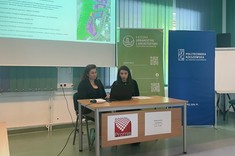 W lekko przyciemnionym pomieszczeniu po środku wysoko zawieszony jasny ekran, przed nim przy stole siedzą dwie prelegentki. W tle kilka wysokich na około 2 metry plansz w zielonym lub niebieskim kolorze z białymi napisami.