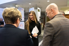 Na zdjęciu młoda kobieta ubrana w czarną sukienkę odbiera nagrodę. Na perwszym planie dwie osoby stojące plecami do fotografa. W tle wystawa: plansze zawieszone na metalowych siatkach, fot. Dawid Zientek