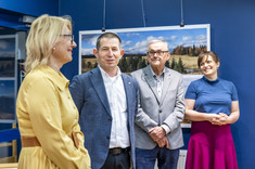 W pomieszczeniu stoją obok siebie cztery osoby, dwóch mężczyzn pośrodku i po bokach kobiety. Rozmawiają ze sobą i uśmiechają się. W tle na granatowej ścianie zawieszone plansze z dużymi kolorowymi fotografiami.