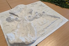 Na jasnym stole leży praca plastyczne. Na kwadratowej podstawie ukształtowane różnej wysokości wzniesienia. Praca jest cała biała.