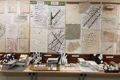 Zbliżenie na ścianę pokrytą modelami miast. Modele w jasnych kolorach. Nieliczne z akcentami kolorystycznymi. Wzdłuż ściany stoją stoły z poustawianymi na nich modelami budynków.